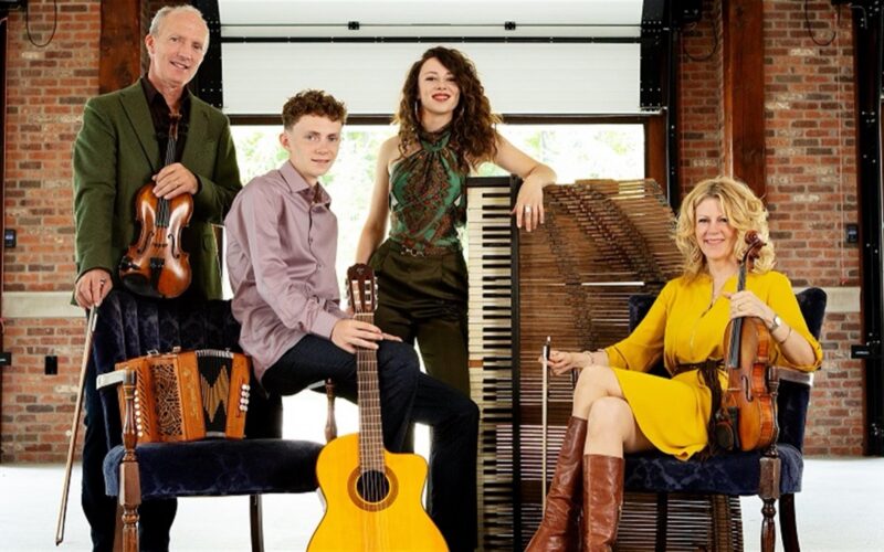 Four musicians in a brick-walled hall with their instruments — two fiddlers, a young man with a guitar, and a young woman beside a piano — the family Celtic ensemble.