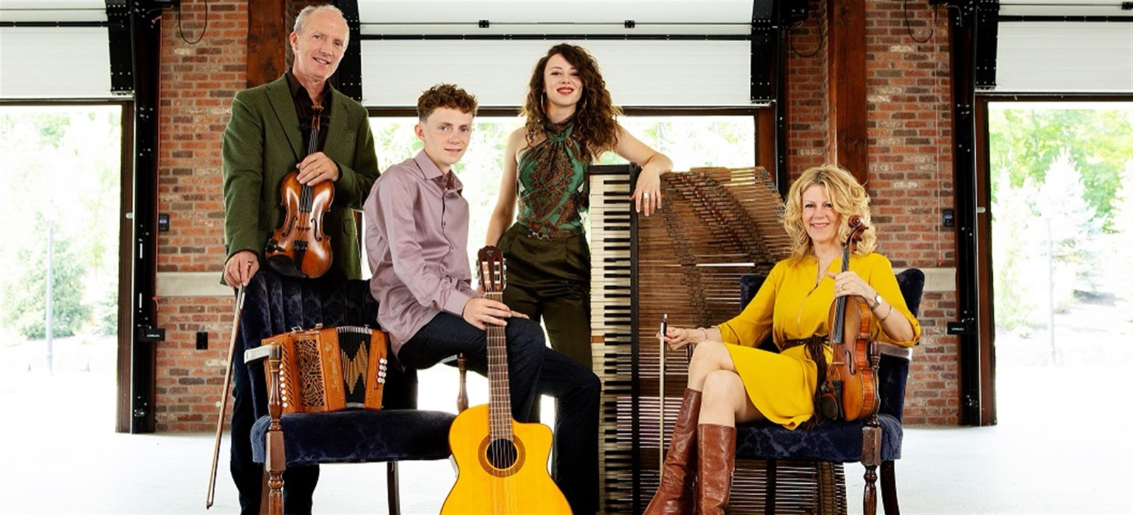 Four musicians in a brick-walled hall with their instruments — two fiddlers, a young man with a guitar, and a young woman beside a piano — the family Celtic ensemble.