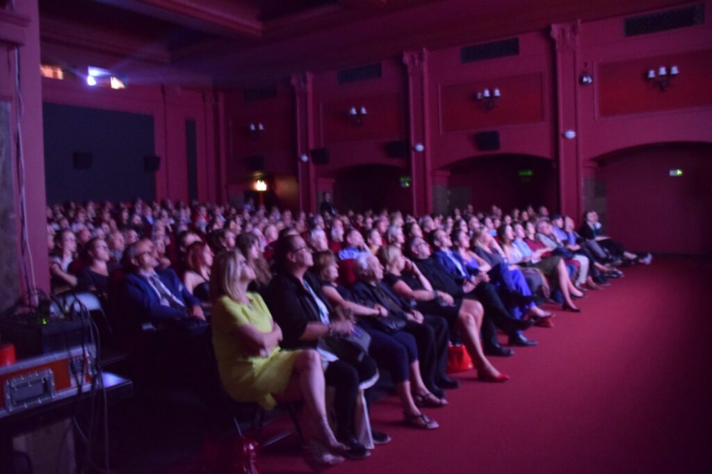 A full audience seated in a red-walled theater, watching the stage intently.