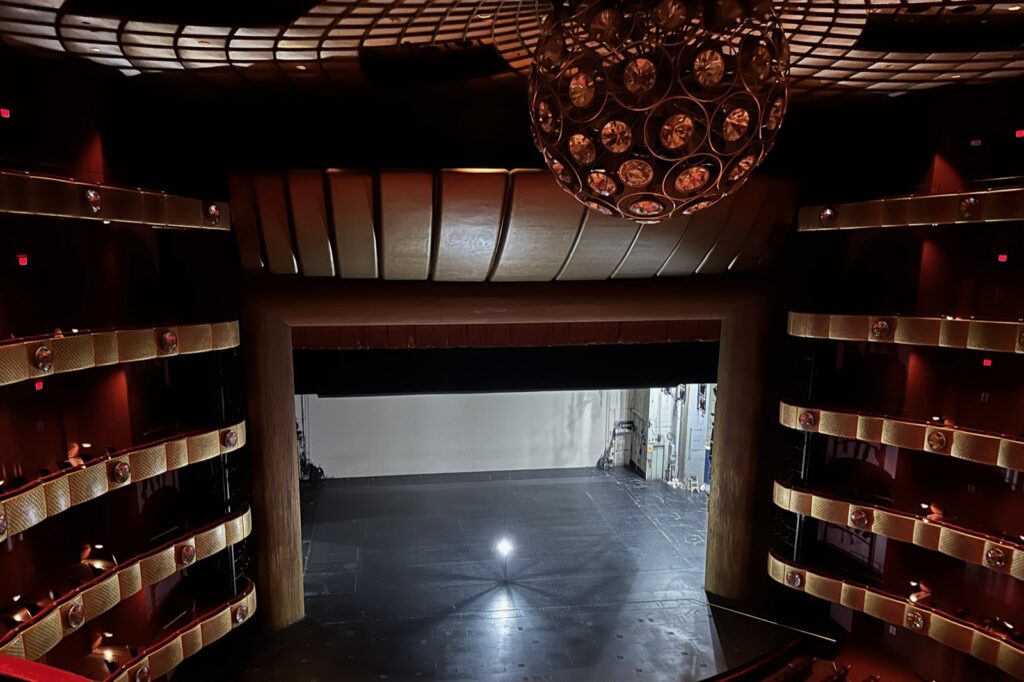 An empty theater auditorium seen from the balcony, its tiers and chandelier in shadow, lit only by a single ghost light standing on the bare stage.