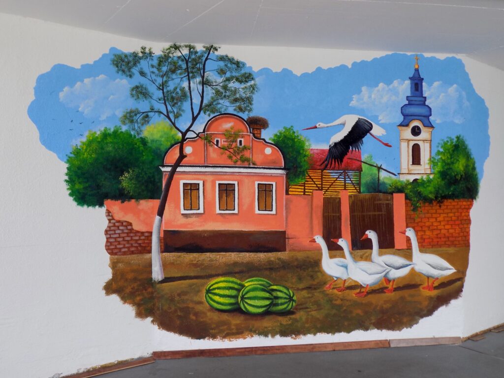 A painted wall mural of a village scene: a pink house with a stork nesting on the chimney, geese in the yard, and watermelons on the ground.
