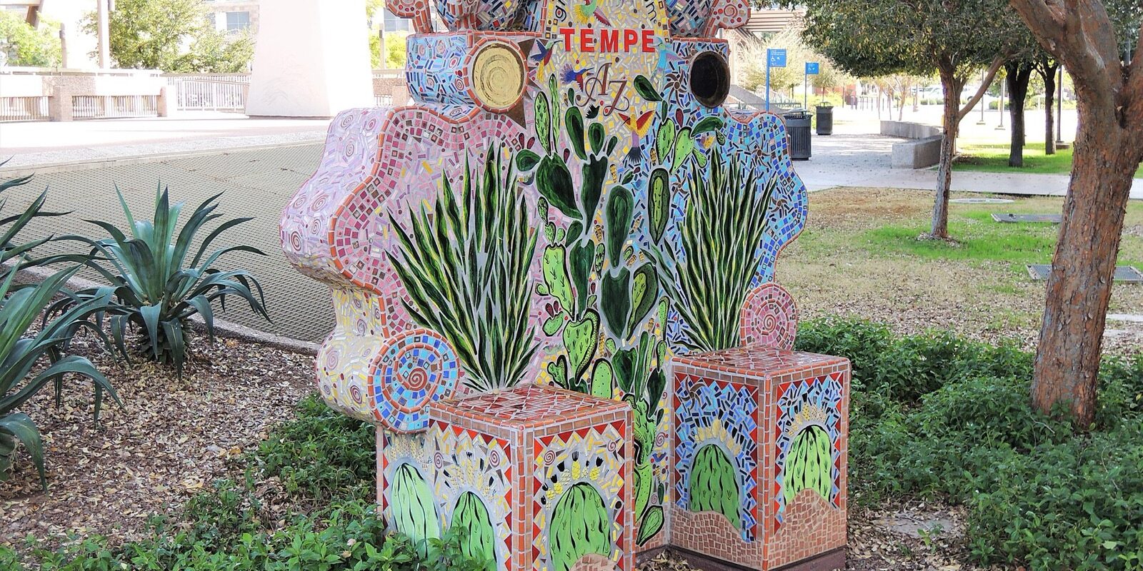 A mosaic-tiled 'Greetings from Tempe AZ' sculpture decorated with cactus and hummingbird motifs, set in a shaded park.